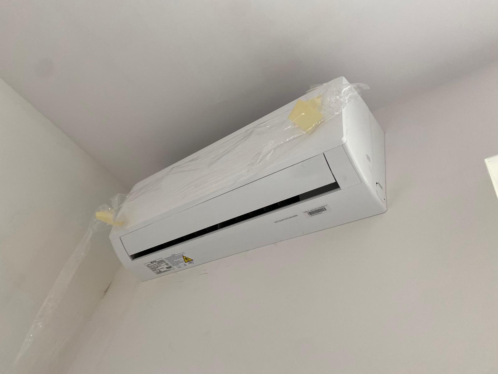 Nowy, biały klimatyzator naścienny, jeszcze w folii ochronnej, zamontowany na białej ścianie w rogu pomieszczenia.