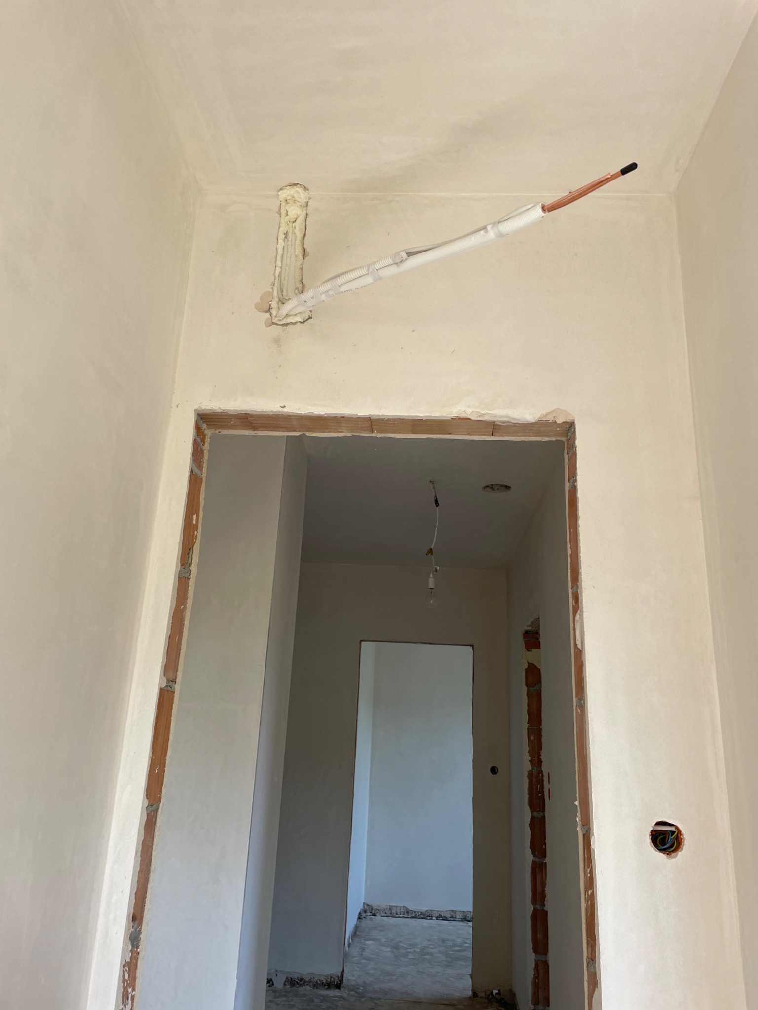 Wewnętrzna instalacja rur miedzianych w izolacji, wystająca ze ściany w surowym wnętrzu z widoczną cegłą wokół framugi drzwi i przewodami elektrycznymi w ścianie.