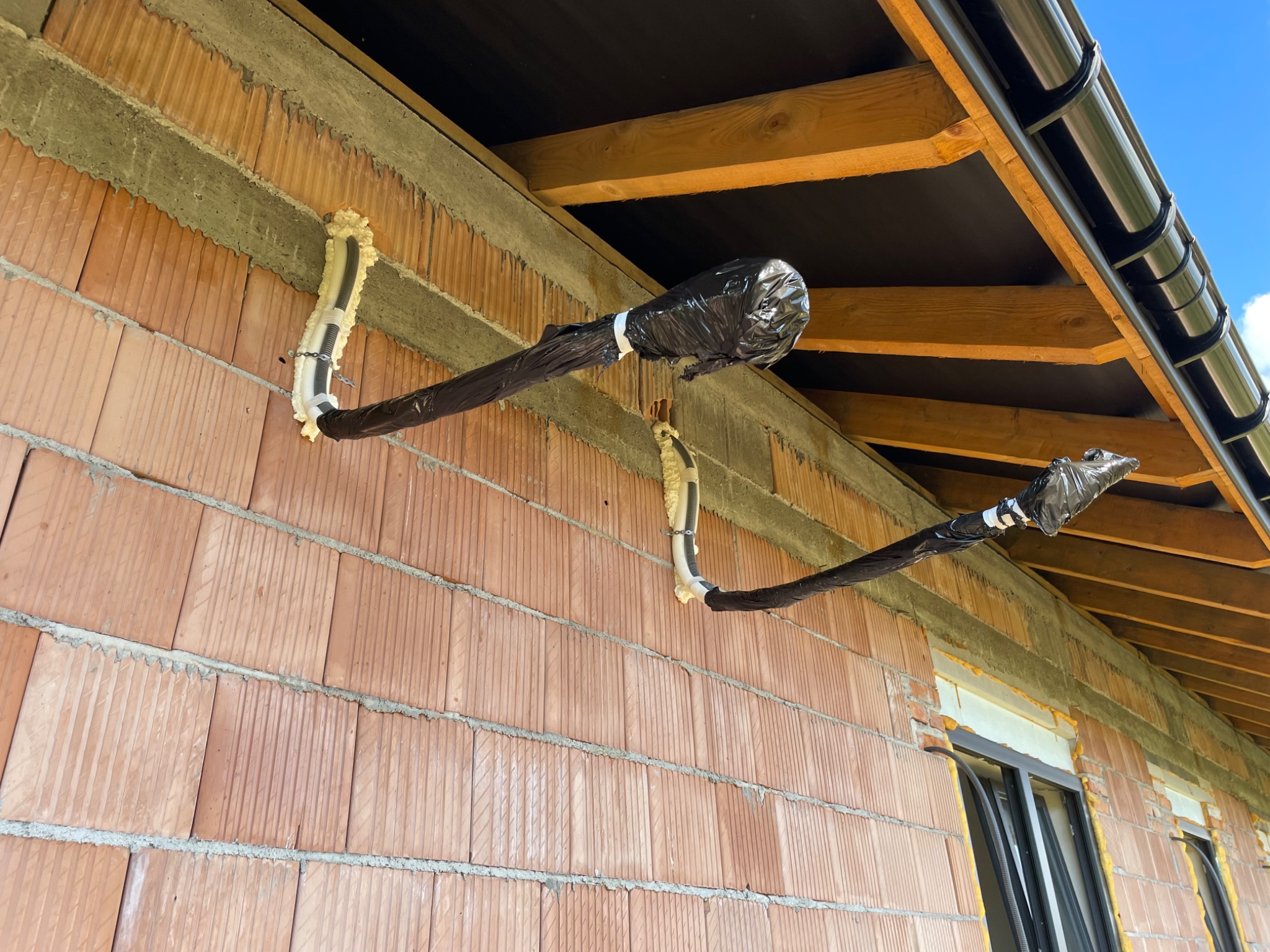 Montaż przewodów klimatyzacji na elewacji z czerwonej cegły, izolowane pianką i czarną folią, widoczne elementy dachu i rynny