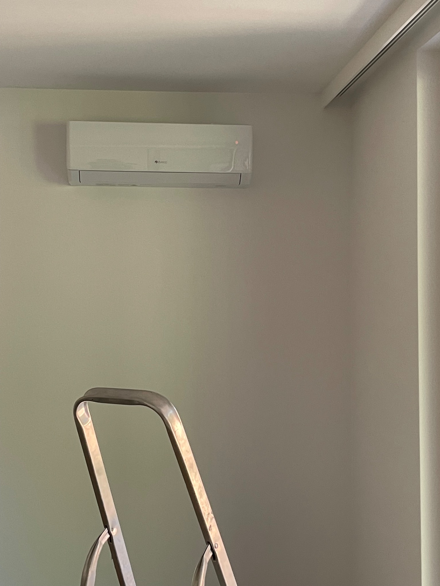 Biała klimatyzacja typu split zamontowana na ścianie nad rozłożoną, metalową drabiną, w pokoju z roletami w kasecie.