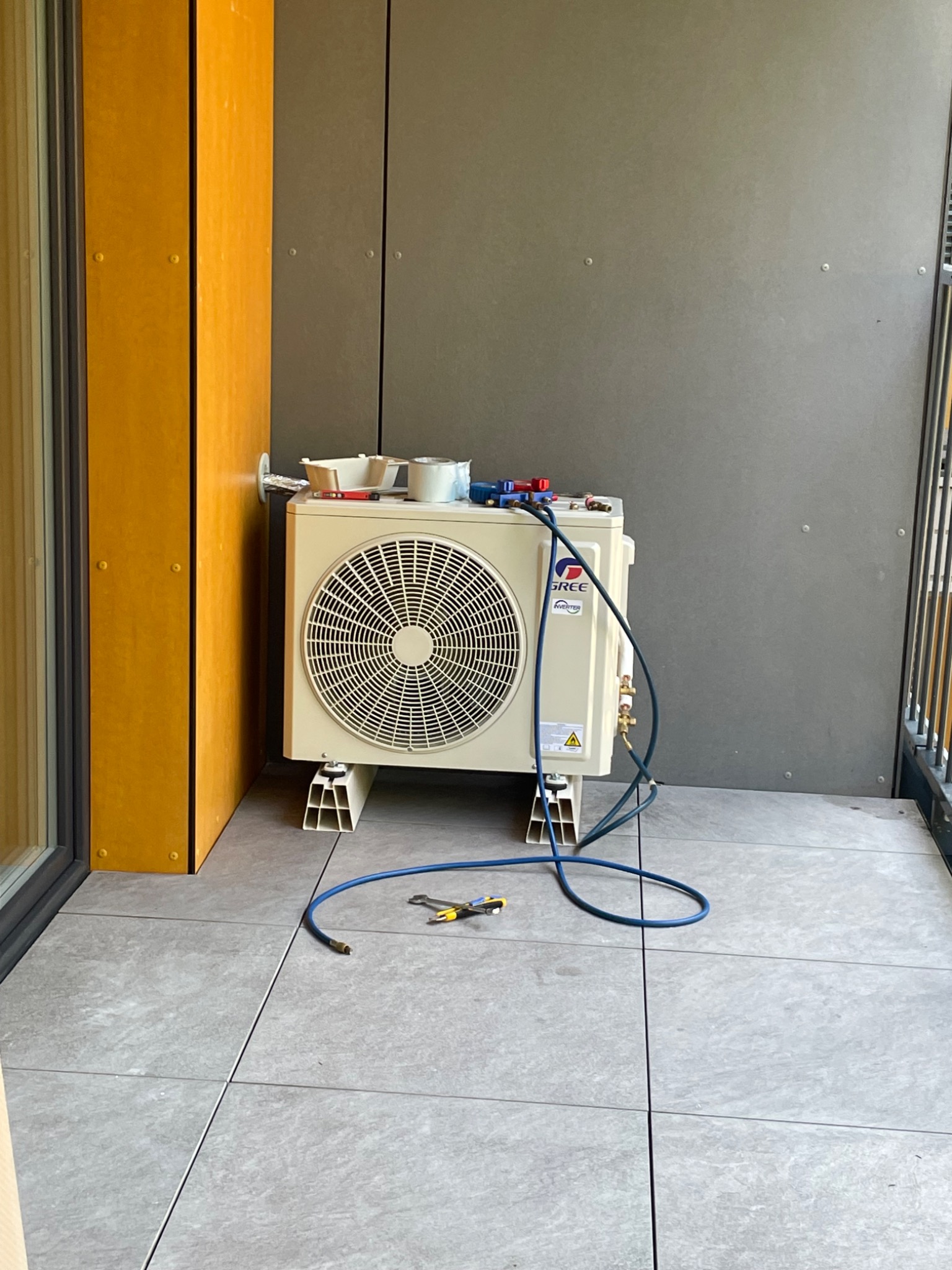 Zewnętrzna jednostka klimatyzacji marki Gree, zamontowana na balkonie na specjalnych wspornikach, z podłączonymi przewodami i narzędziami serwisowymi na górze, widoczne płytki podłogowe i fragment...