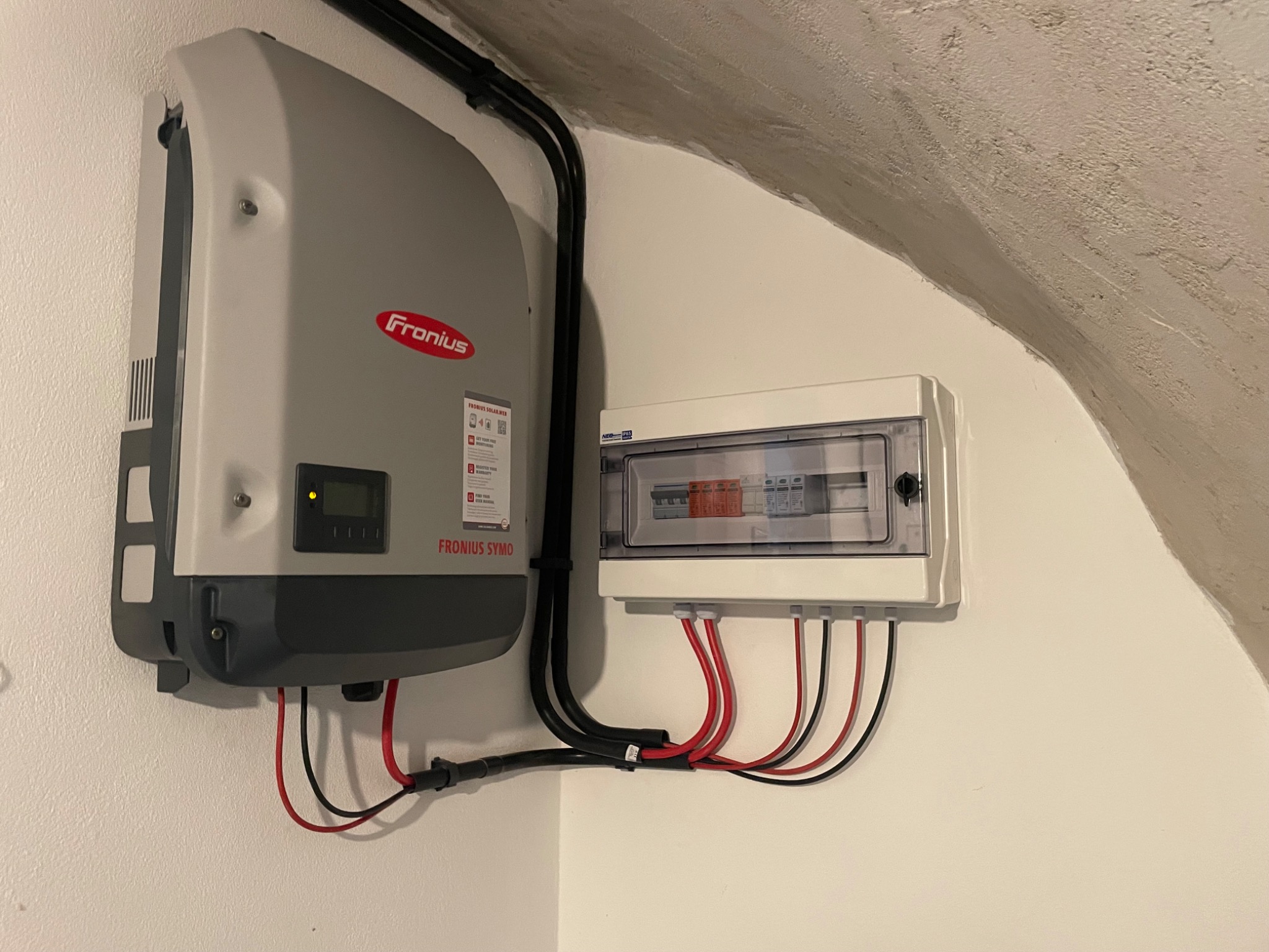 Szary falownik Fronius Symo z podłączonymi kablami oraz skrzynka z bezpiecznikami zamontowane na białej ścianie w Krakowie, widoczne czarne kable w peszlu.