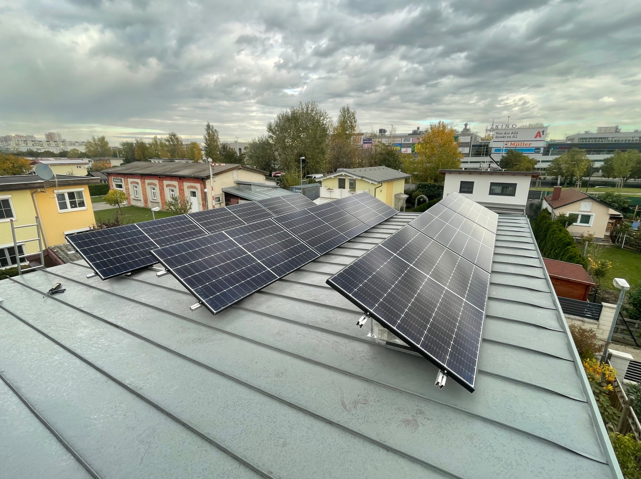 Panele słoneczne zamontowane na szarym metalowym dachu budynku mieszkalnego w pochmurny dzień, z widokiem na zabudowę miejską w tle.