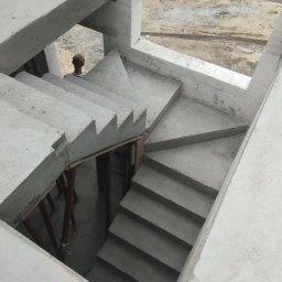 OlegDyshko - Surowe, betonowe schody w trakcie budowy domu murowanego, widoczne białe bloczki i podpory schodów, w tle sylwetka osoby.