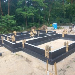 OlegDyshko - Zabezpieczone, pomalowane na czarno fundamenty domu jednorodzinnego, z widocznymi drewnianymi stemplami szalunkowymi, na piaszczystym podłożu, w tle las i przenośna toaleta.