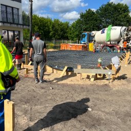 OlegDyshko - Wylewanie betonu na przygotowane zbrojenie fundamentu domu jednorodzinnego, widoczny betonowóz z pompą, szalunek z desek i pracownicy budowlani w trakcie pracy.