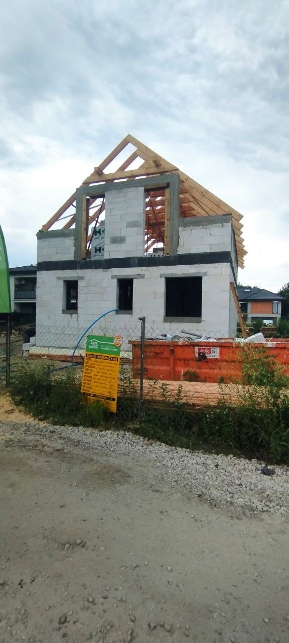 Surowy stan domu z białych bloczków z drewnianą konstrukcją dachu, widoczny kontener budowlany i tablica informacyjna budowy.