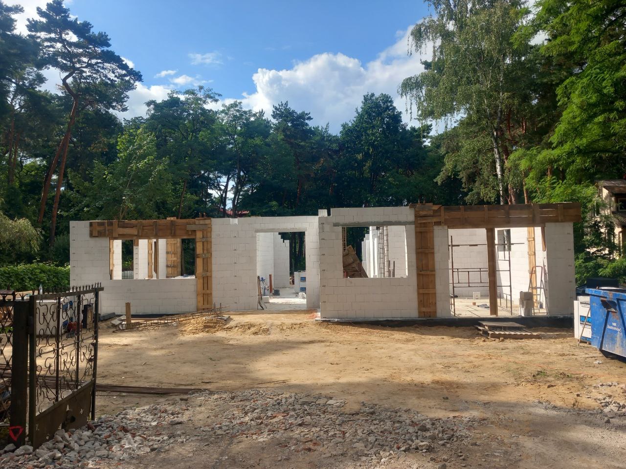 Wznoszenie ścian parteru domu z bloczków betonu komórkowego, z widocznym szalunkiem drewnianym nad otworami okiennymi, na tle zieleni drzew i błękitnego nieba.