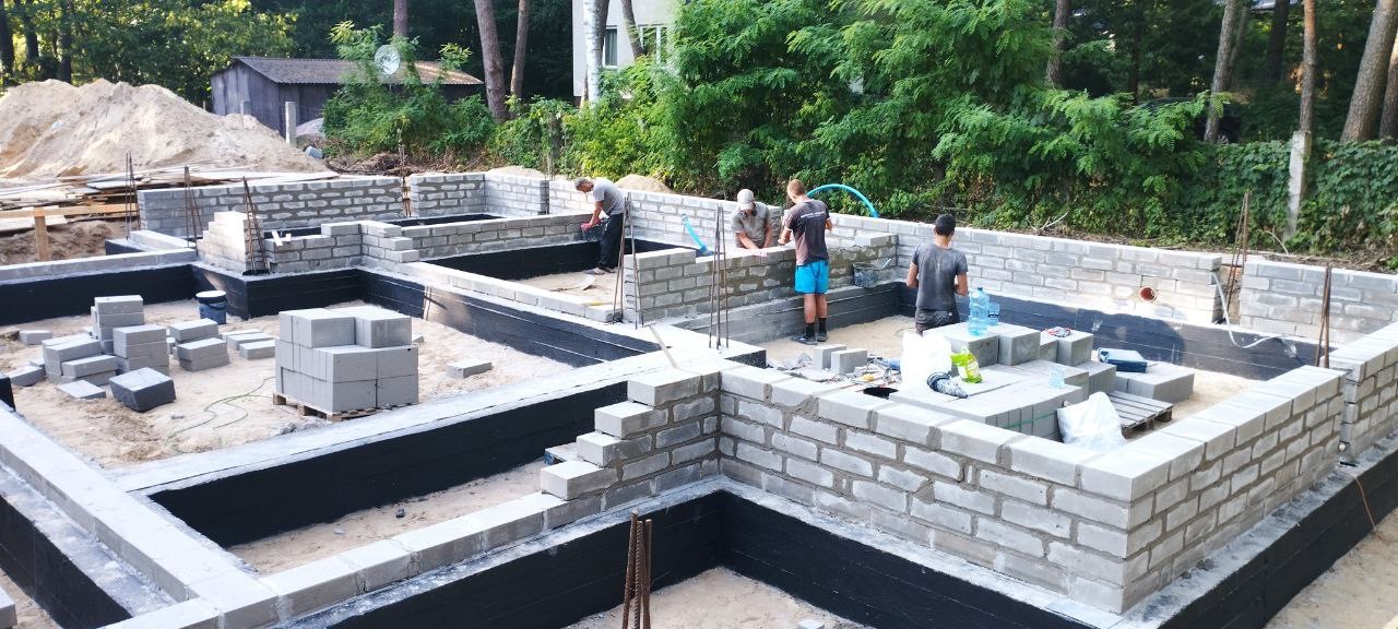 Budowa fundamentów z bloczków betonowych, widoczna izolacja przeciwwilgociowa, pracownicy murują ściany fundamentowe, w tle drzewa i budynek gospodarczy.