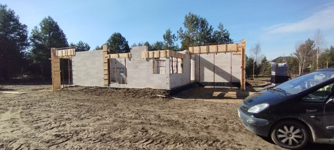Surowy stan ścian z betonu komórkowego w trakcie budowy domu, widoczne drewniane szalunki okienne i drzwiowe, czarna izolacja przeciwwilgociowa fundamentów, w tle las i częściowo widoczny samochód...