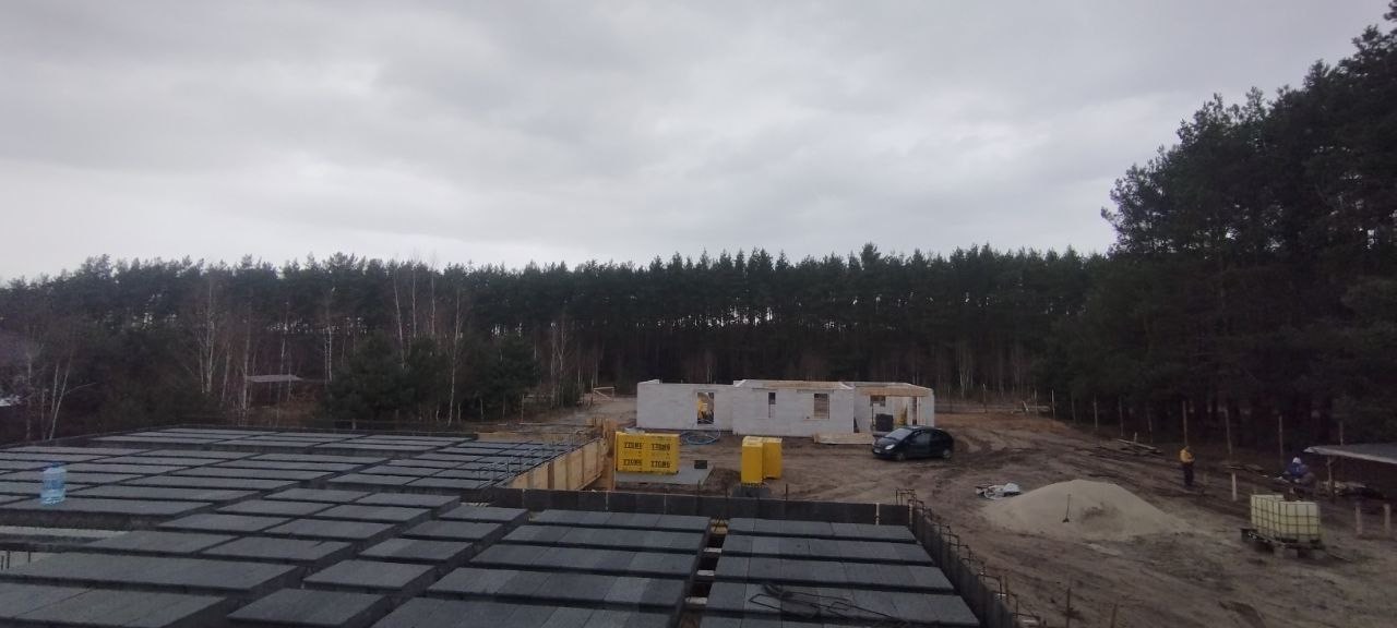 Budowa domu murowanego w stanie surowym otwartym z widocznym stropem Teriva, bloczkami Ytong i lasem w tle, pochmurny dzień.