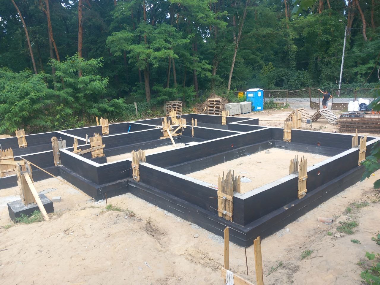 Zabezpieczone, pomalowane na czarno fundamenty domu jednorodzinnego, z widocznymi drewnianymi stemplami szalunkowymi, na piaszczystym podłożu, w tle las i przenośna toaleta.