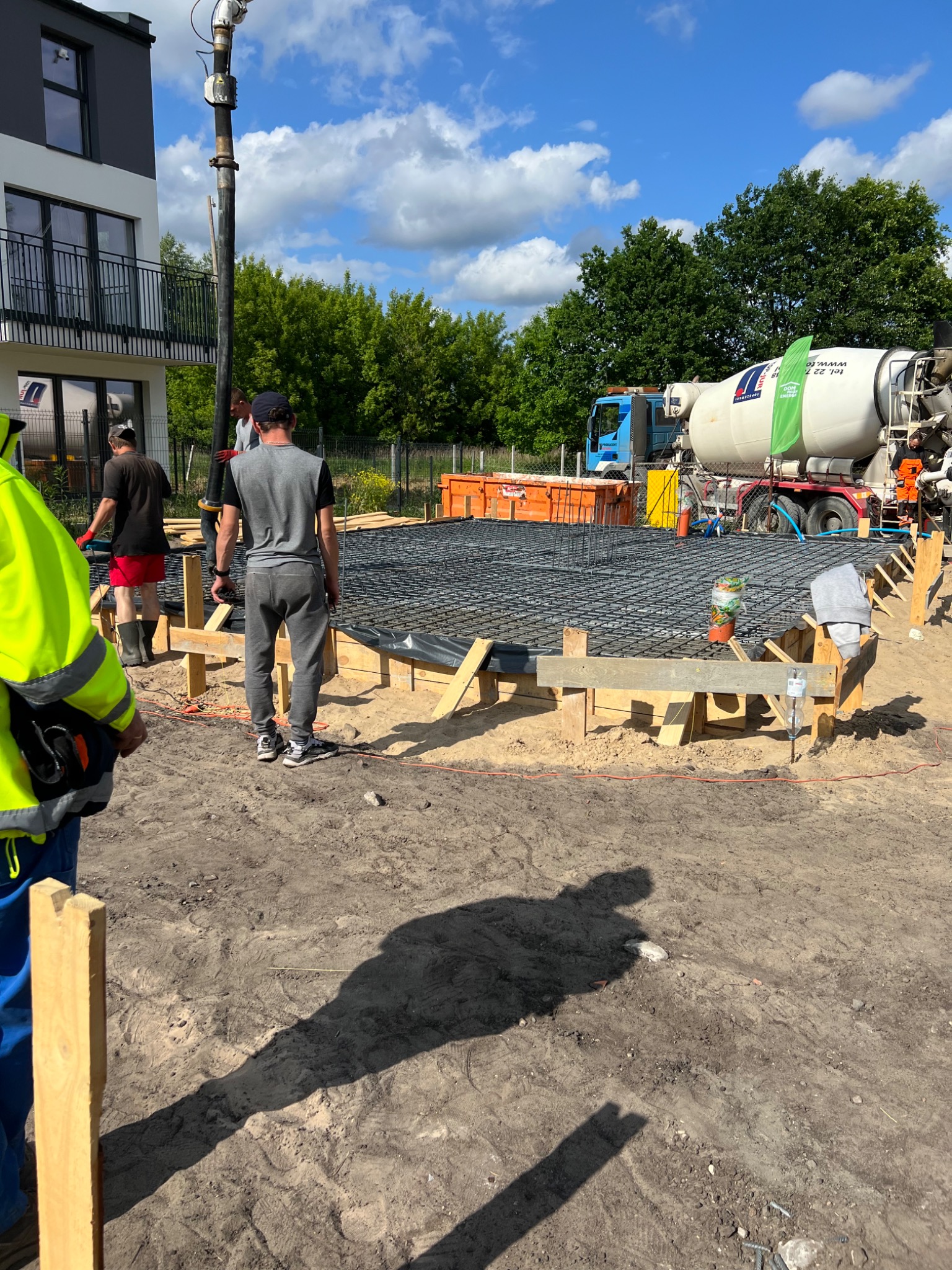 Wylewanie betonu na przygotowane zbrojenie fundamentu domu jednorodzinnego, widoczny betonowóz z pompą, szalunek z desek i pracownicy budowlani w trakcie pracy.