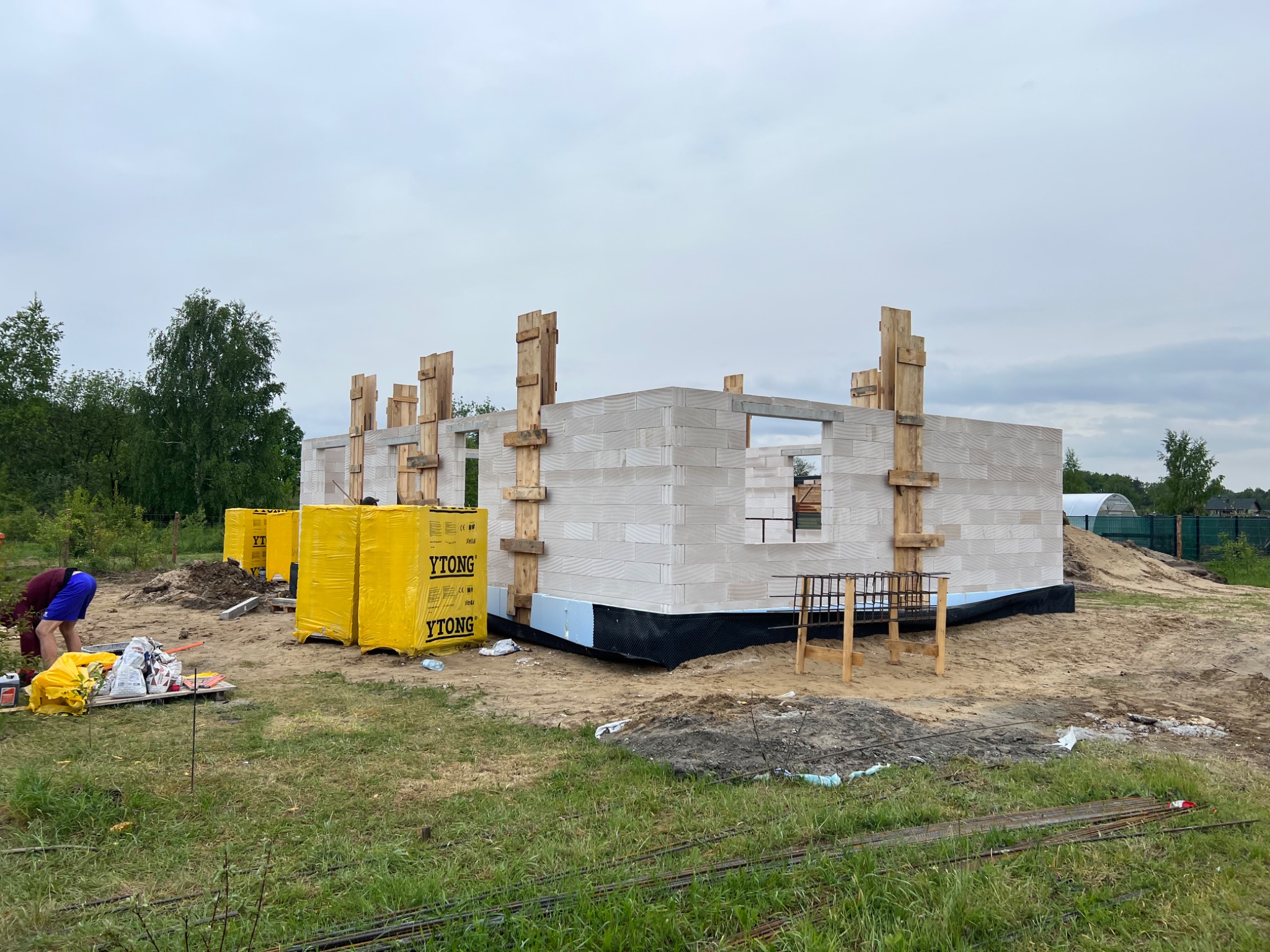 Wznoszenie ścian parterowego domu z bloczków Ytong, widoczne drewniane stemple podtrzymujące, zabezpieczenie fundamentów folią kubełkową, palety z bloczkami na trawiastym terenie budowy.