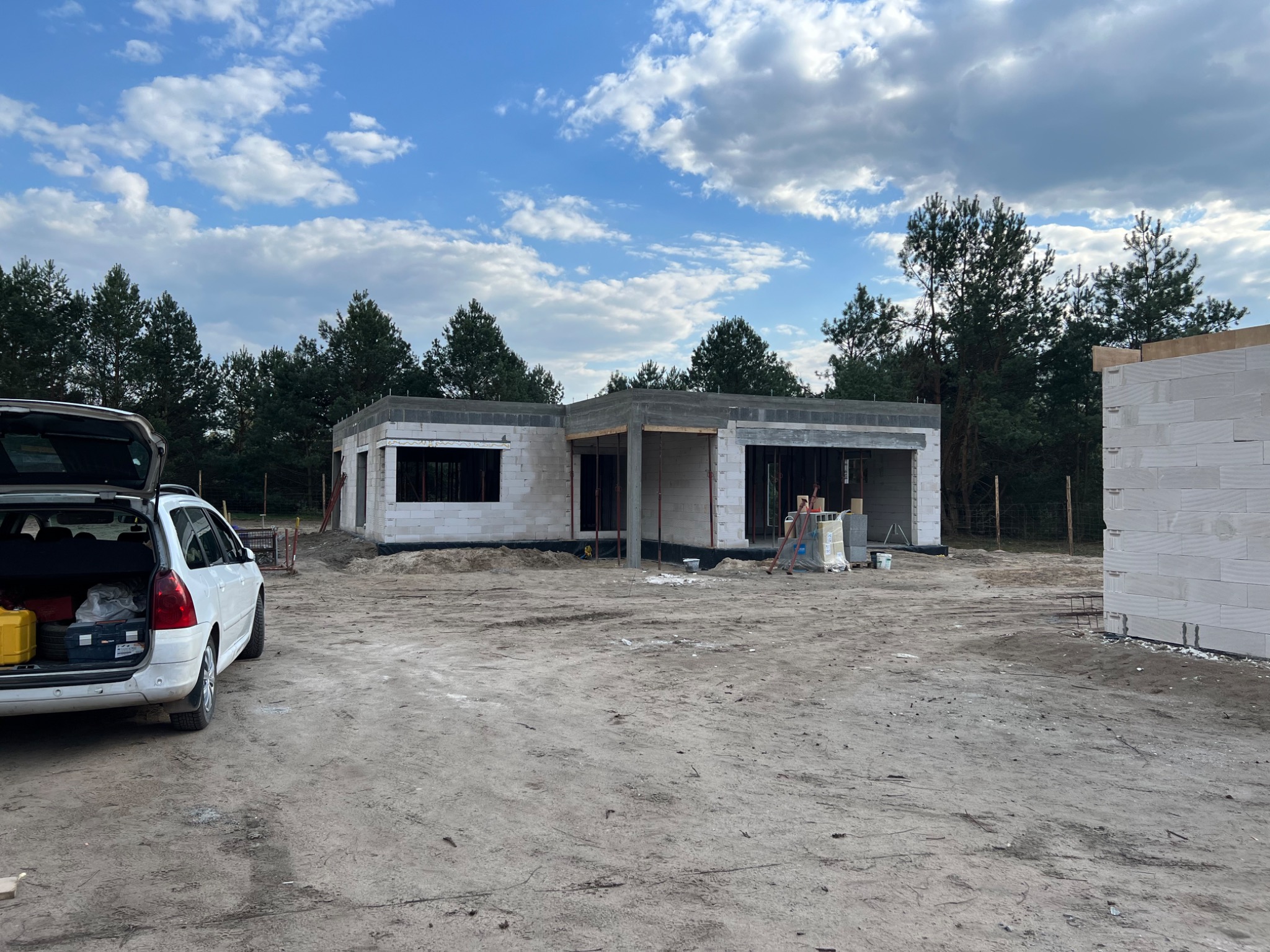 Surowy stan domu z białych bloczków, widoczny z zewnątrz, z otwartym bagażnikiem samochodu osobowego po lewej stronie i lasem w tle.