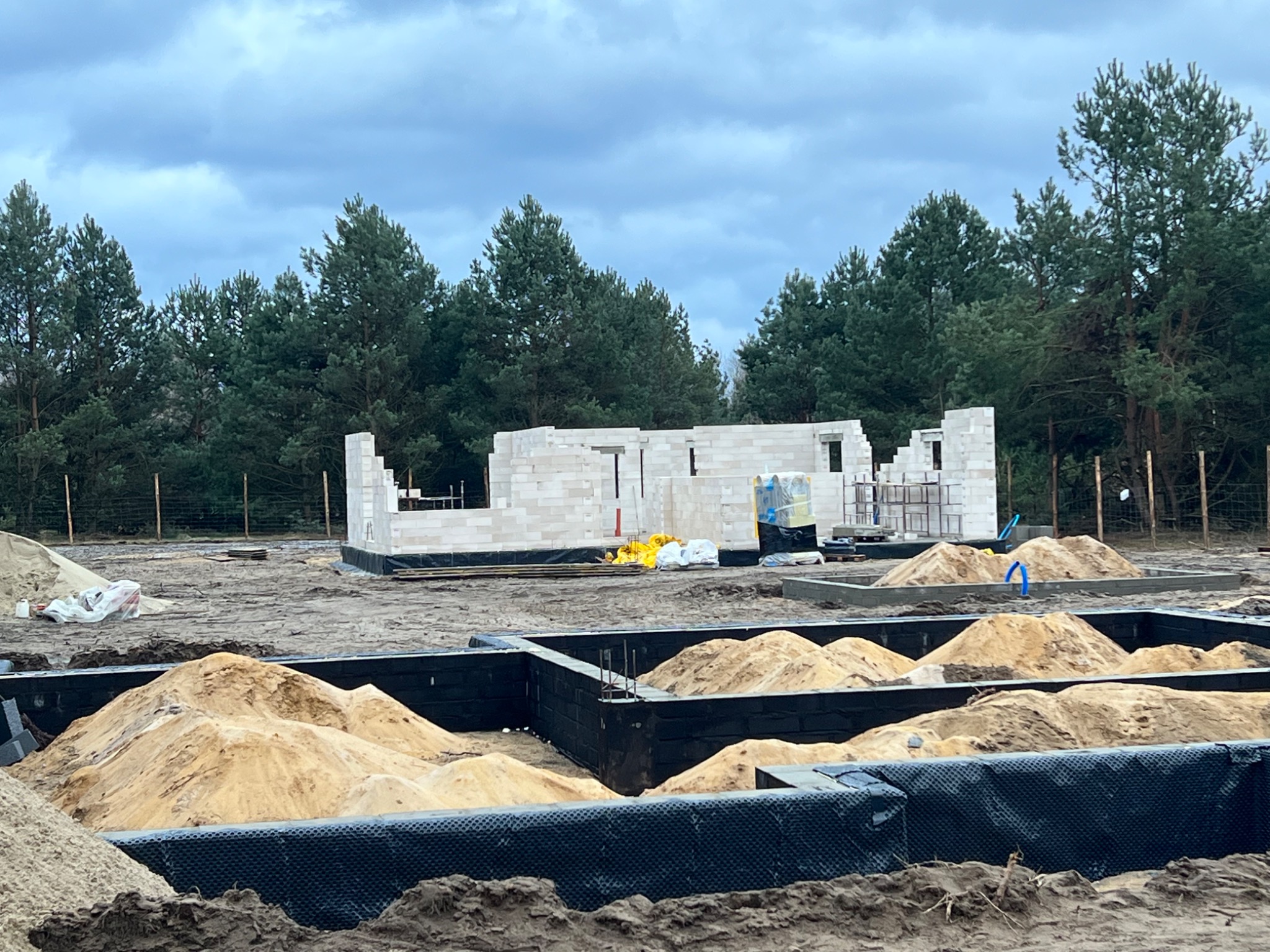 Budowa domu murowanego: widok na fundamenty z izolacją i początkowe ściany z białych bloczków, w tle las iglasty i pochmurne niebo.