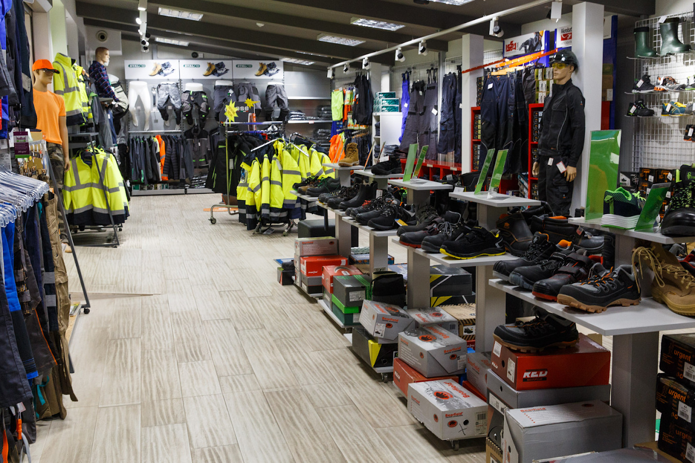 Wnętrze sklepu z odzieżą i obuwiem roboczym, z szerokim wyborem kurtek ostrzegawczych, spodni, kombinezonów i butów na półkach i wieszakach.