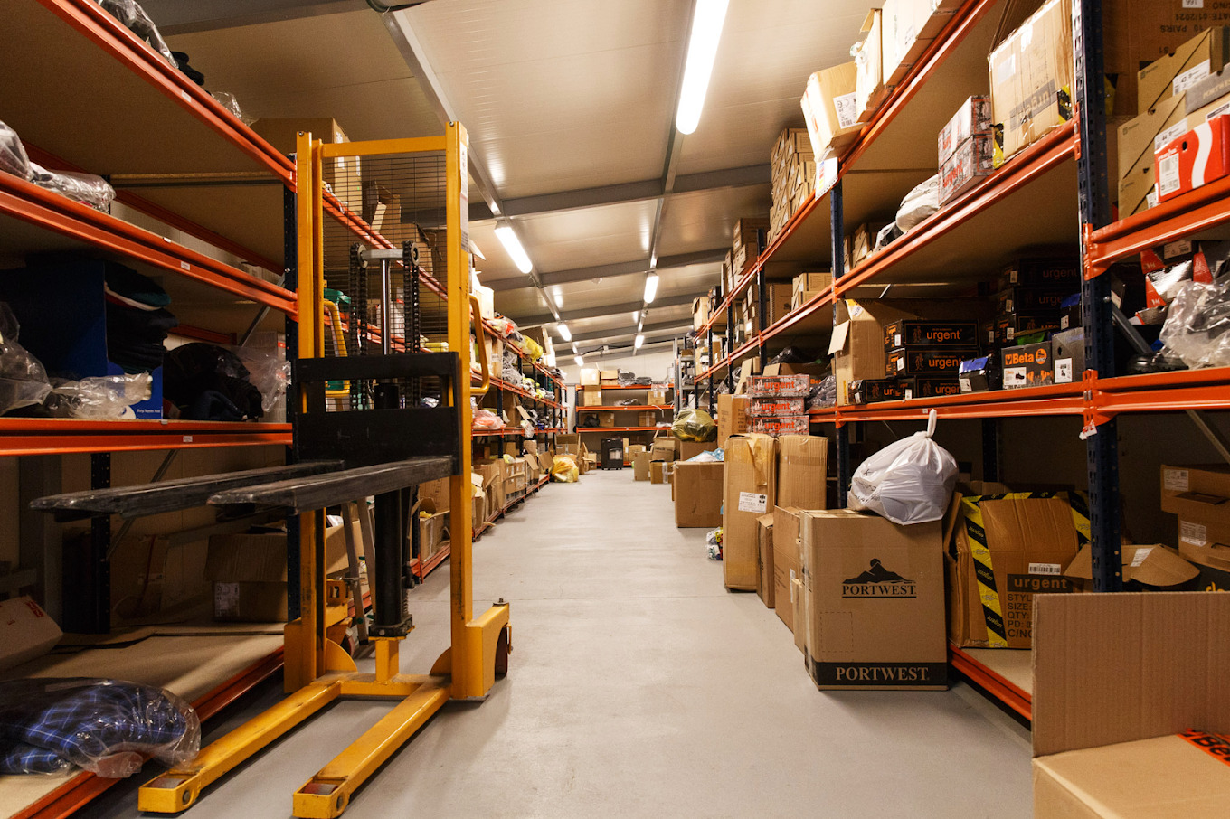 Magazyn z regałami wypełnionymi kartonami i odzieżą, widoczny wózek widłowy w kolorze żółtym.