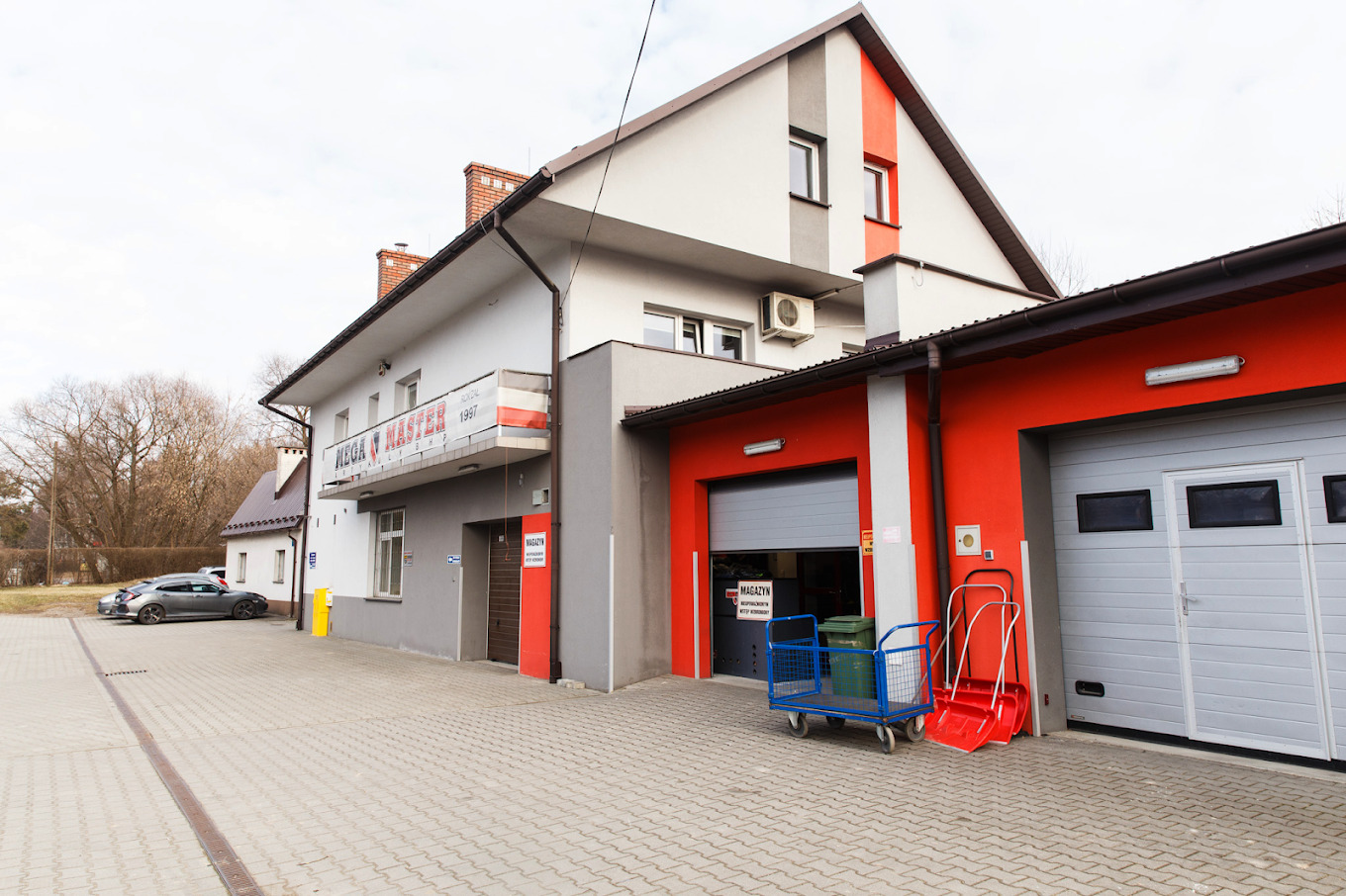 Budynek firmy odzieżowej z widocznym magazynem, wózkiem transportowym i szuflami do śniegu przed garażem.