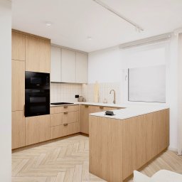 Cholewa - Interior Design Concept - Jasna kuchnia z drewnianymi frontami, czarnym piekarnikiem i mikrofalówką w zabudowie. Na blacie złota bateria i akcesoria. Parkiet w jodełkę na podłodze.