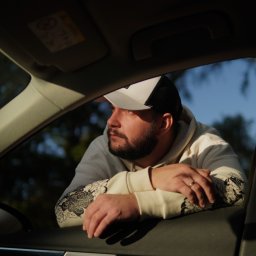 Andrii Shestakov - Mężczyzna w czapce i bluzie z wzorem, oparty o otwarte okno samochodu, patrzy w dal. Widoczne wnętrze auta i fragment nieba z drzewami w tle.