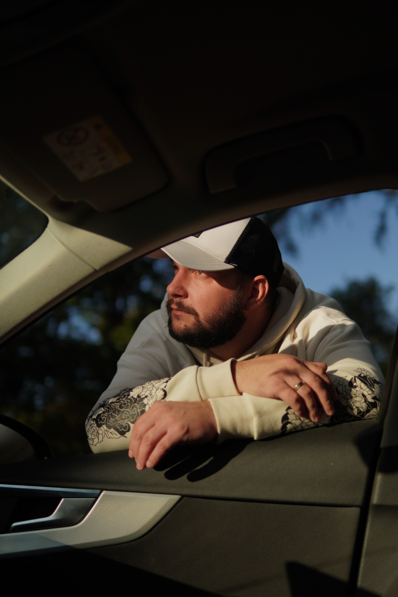 Mężczyzna w czapce i bluzie z wzorem, oparty o otwarte okno samochodu, patrzy w dal. Widoczne wnętrze auta i fragment nieba z drzewami w tle.