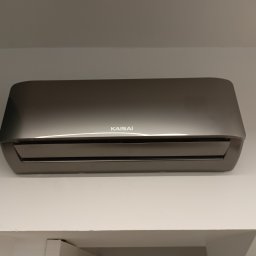 ML- Marcin Łowczyński - Szary klimatyzator Kaisai zamontowany na białej ścianie, widok od dołu, fragment sufitu widoczny w kadrze.