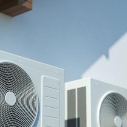 ML- Marcin Łowczyński - Dwa białe klimatyzatory zamontowane na elewacji budynku, z widocznym balkonem w tle i fragmentem zieleni.