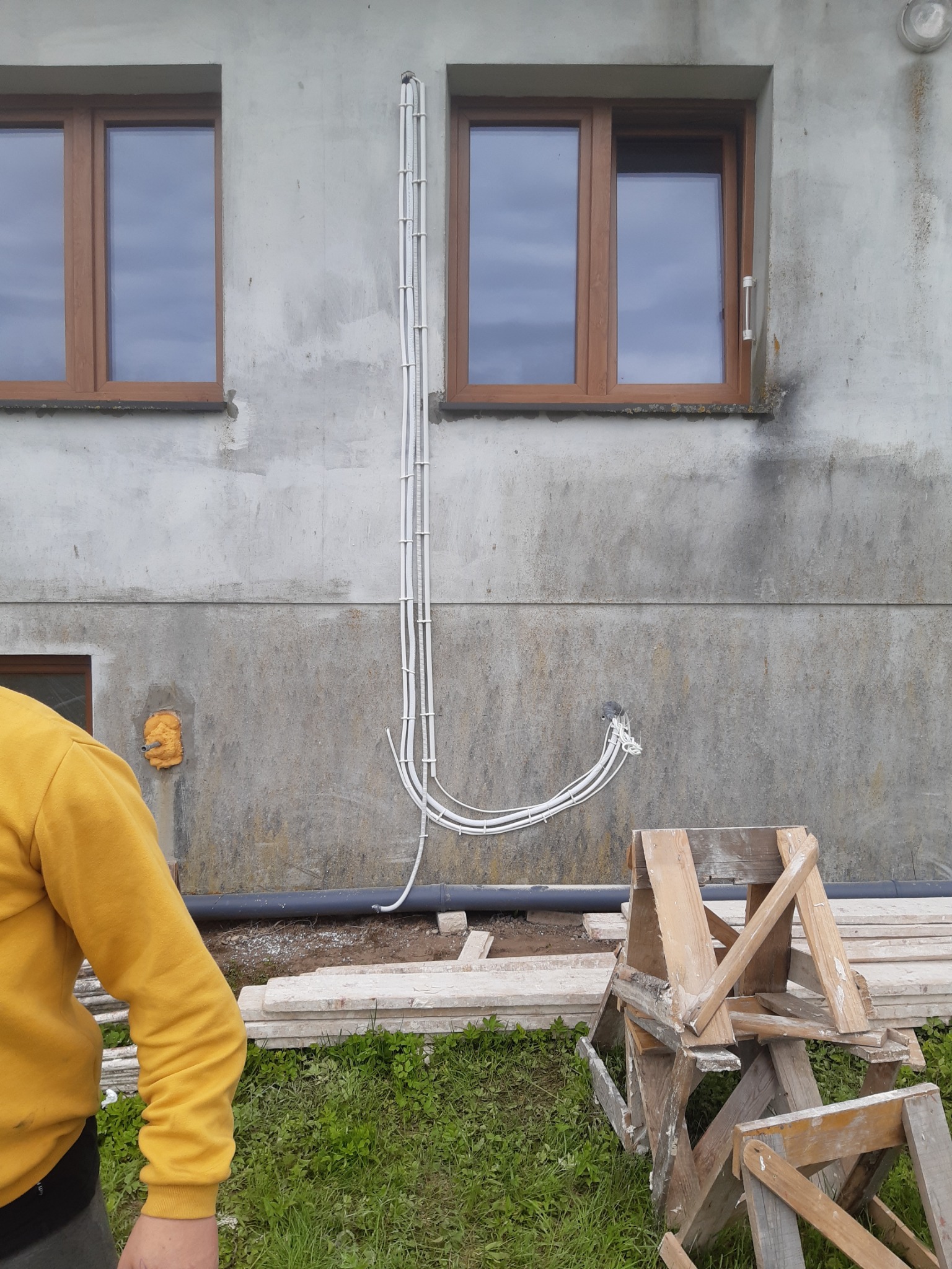 Białe kable elektryczne ułożone wzdłuż szarej ściany zewnętrznej budynku z brązowymi oknami, widoczne drewniane rusztowanie i fragment osoby w żółtej bluzie.