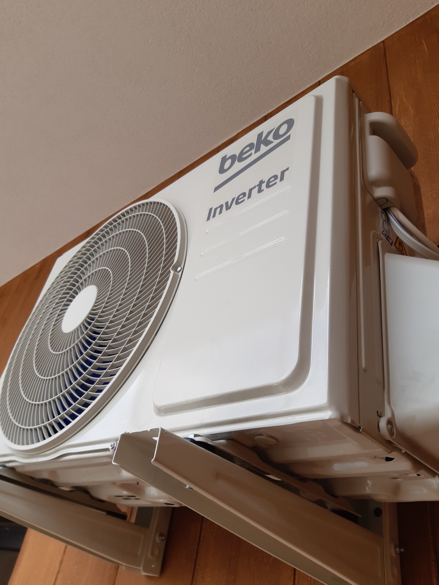 Biała jednostka wewnętrzna klimatyzacji Beko Inverter zamontowana na drewnianej ścianie, widok z dołu, widoczne przewody.