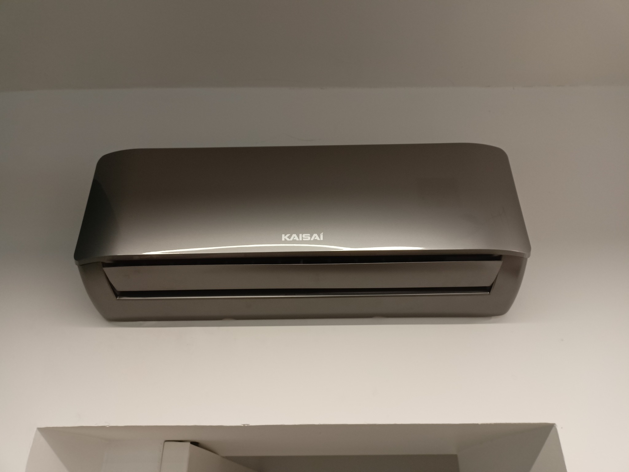 Szary klimatyzator Kaisai zamontowany na białej ścianie, widok od dołu, fragment sufitu widoczny w kadrze.