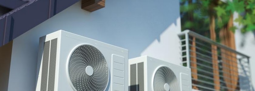 Dwa białe klimatyzatory zamontowane na elewacji budynku, z widocznym balkonem w tle i fragmentem zieleni.