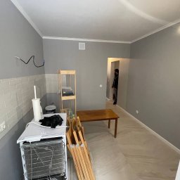 DADAEVS SPÓŁKA Z OGRANICZONĄ ODPOWIEDZIALNOŚCIĄ - Wnętrze pokoju w trakcie odnawiania: szare ściany, częściowo wyłożone białymi płytkami imitującymi cegłę, widoczne przewody elektryczne, drewniany stół i regał, biała lodówka turystyczna oraz w tle...