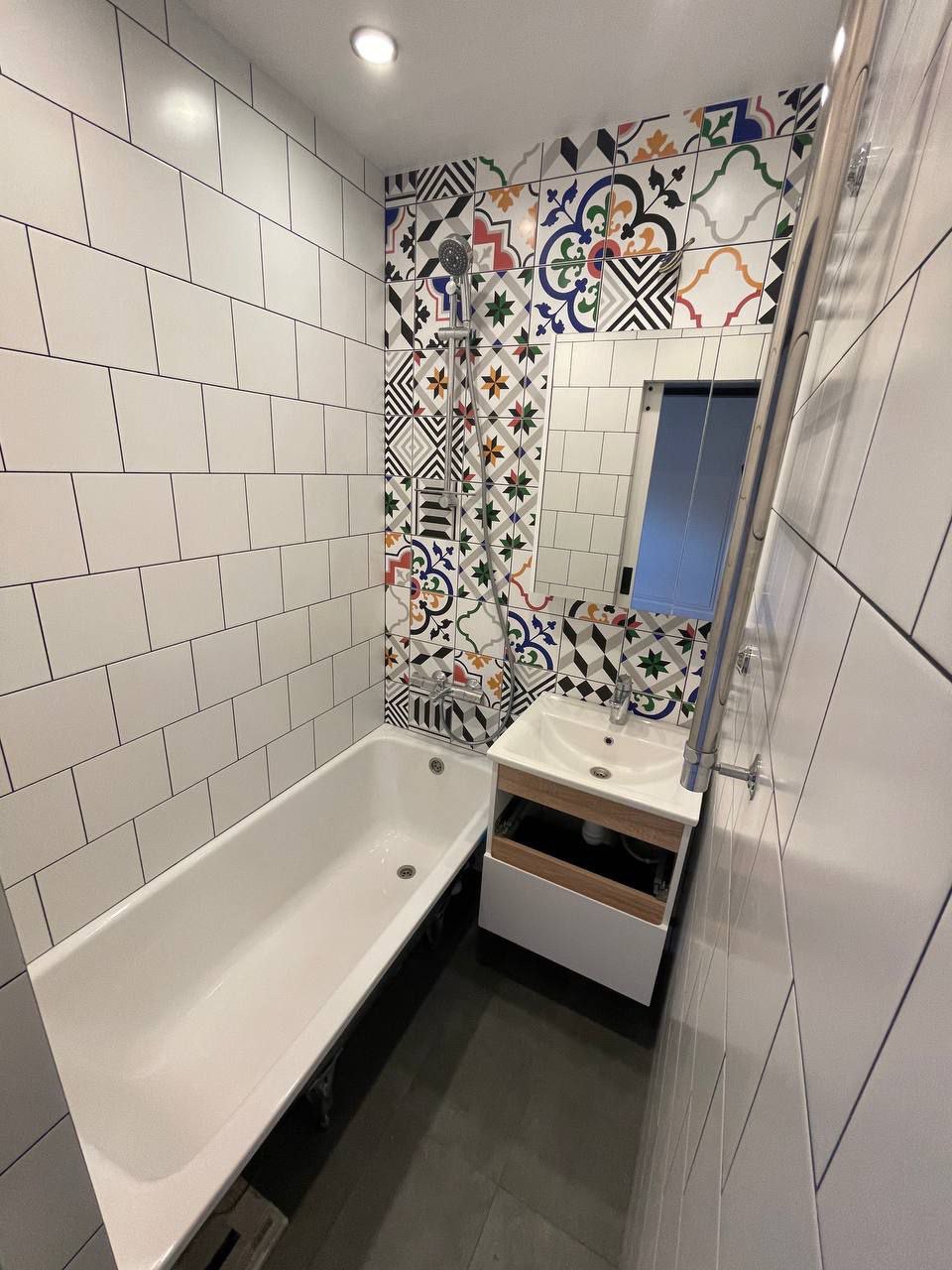 Wykończona łazienka z białą wanną, umywalką z szafką w kolorze drewna i białym blatem oraz ścianą z białych płytek i dekoracyjnych płytek patchworkowych nad wanną.