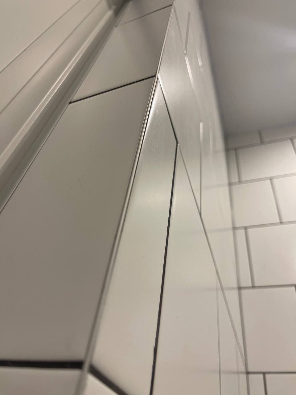 Fragment wykończonej ściany łazienki z białymi, prostokątnymi płytkami ułożonymi w cegiełkę, widoczne fugi w kolorze czarnym, narożnik wykończony listwą.