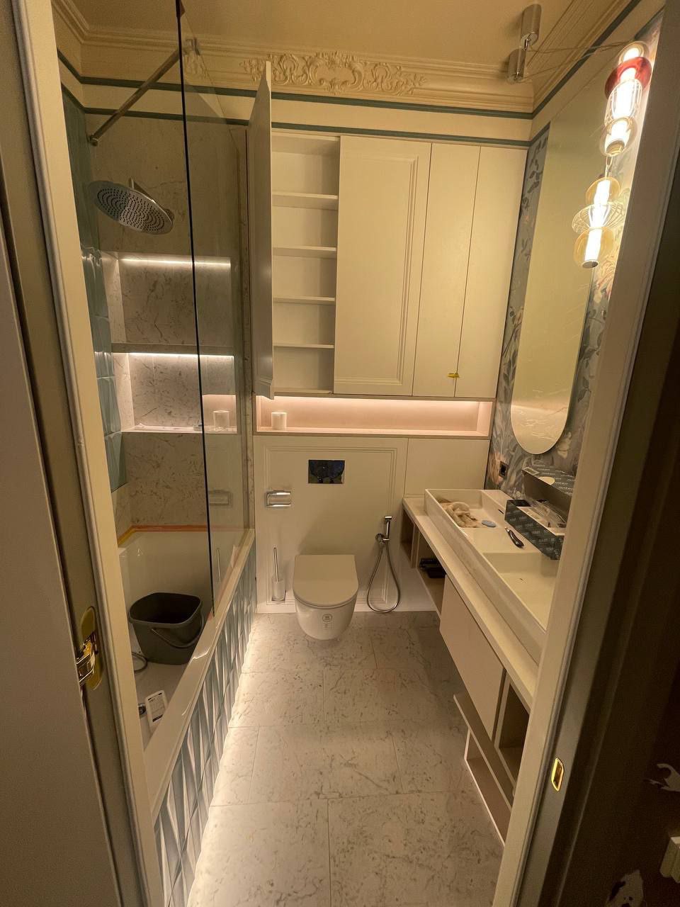 Nowoczesna łazienka z wanną za szklaną ścianką, wiszącą toaletą, umywalką z szafką oraz szafką wiszącą z otwartymi półkami, oświetlona listwami LED i ozdobną lampą.