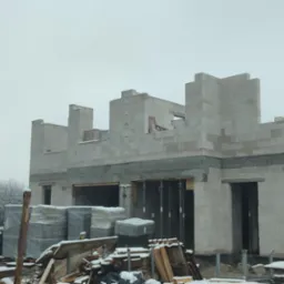 Surowy stan domu w trakcie budowy z białych bloczków, widoczne otwory okienne i drzwiowe, na pierwszym planie palety z materiałami budowlanymi pod śniegiem, w tle biały samochód dostawczy...