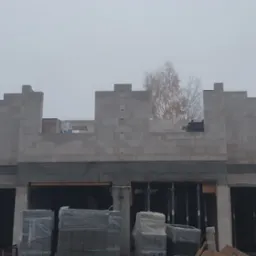 Surowy stan domu z betonu komórkowego w trakcie budowy, widoczne palety z materiałem budowlanym i zarys okien w szary, mglisty dzień.