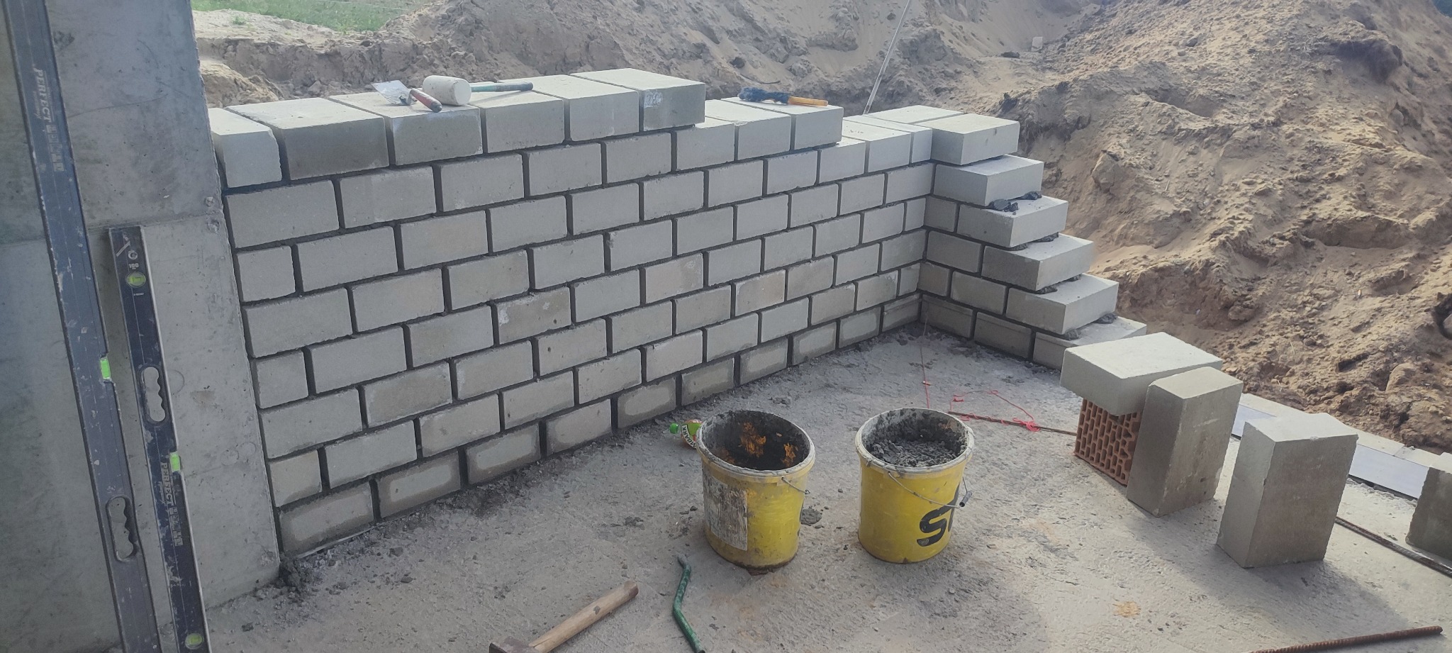 Wznoszona ściana z szarych bloczków, widoczne dwa żółte wiadra z zaprawą, poziomica oparta o ścianę, w tle usypana ziemia.