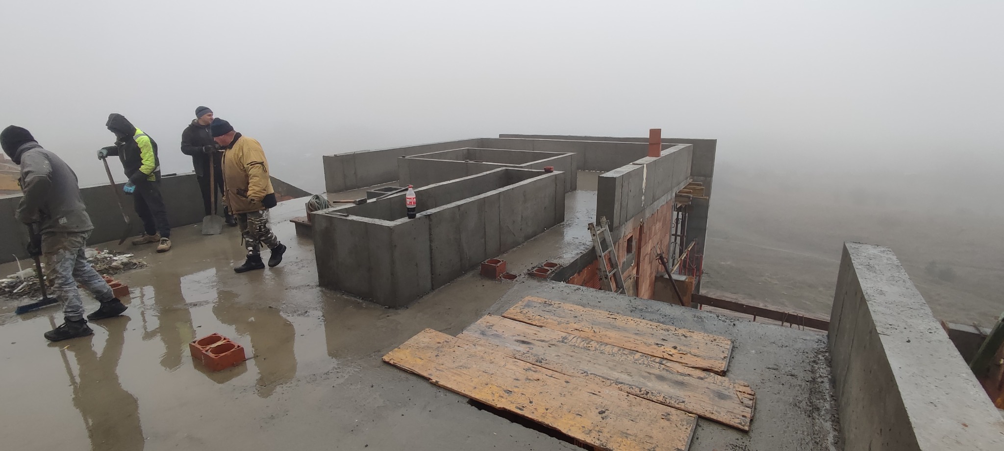 Budowa domu w trakcie wznoszenia ścian fundamentowych i pierwszych warstw muru, widoczni robotnicy z łopatami, butelka Coca-Coli na murze, mglista pogoda.