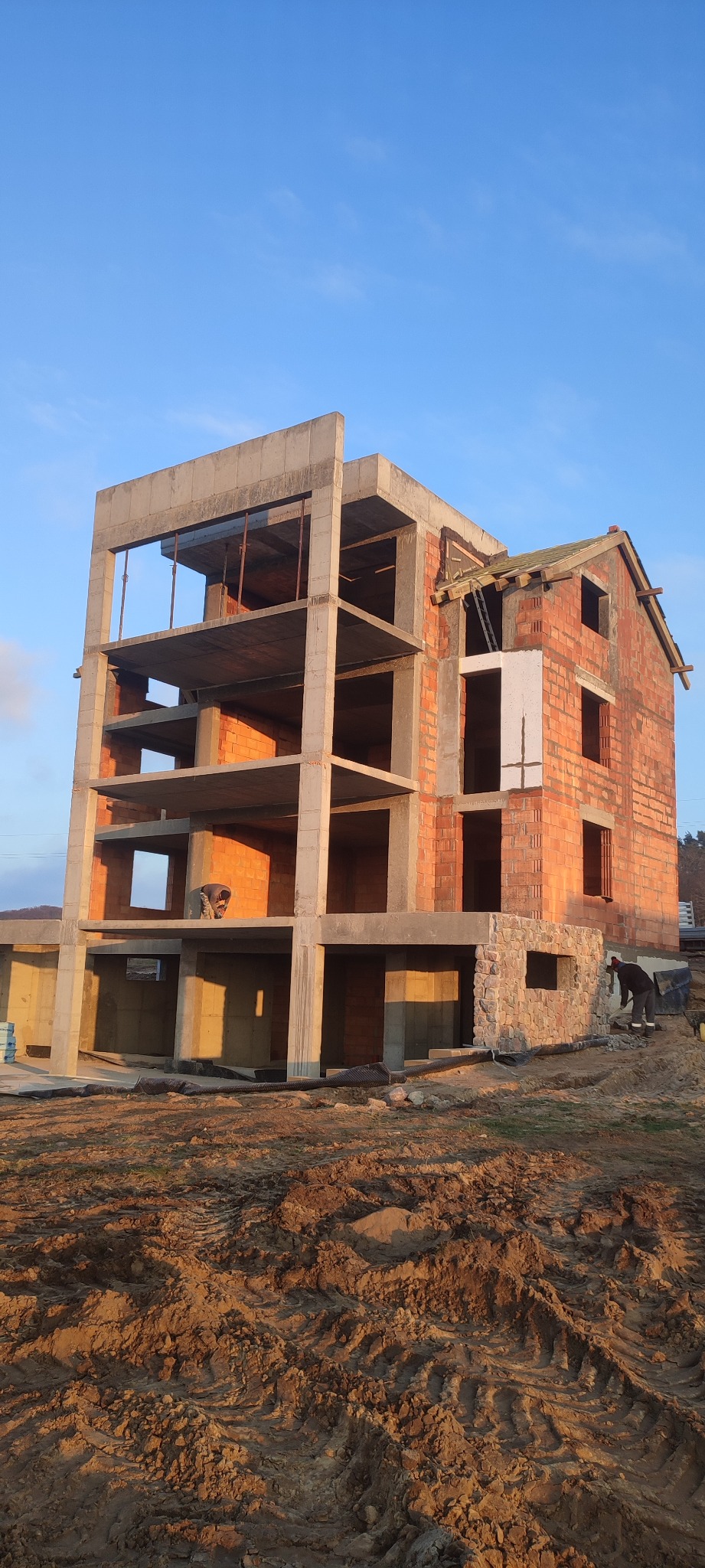 Surowy stan budynku w trakcie budowy, z widocznymi elementami konstrukcji żelbetowej i murowanymi ścianami z cegły, prace ziemne w tle.