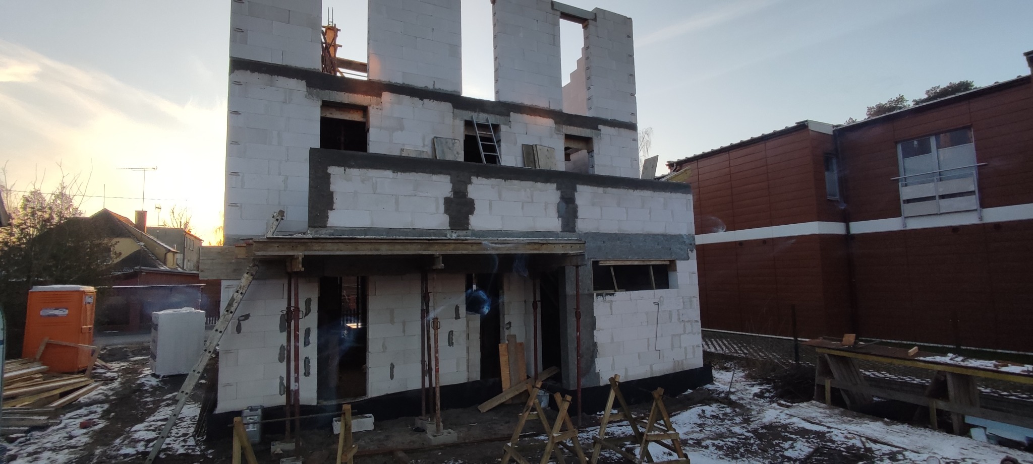 Wznoszenie dwukondygnacyjnego domu z białych bloczków, widoczne szare nadproża, podpierające stemple i drewniane kozły na ośnieżonym terenie.