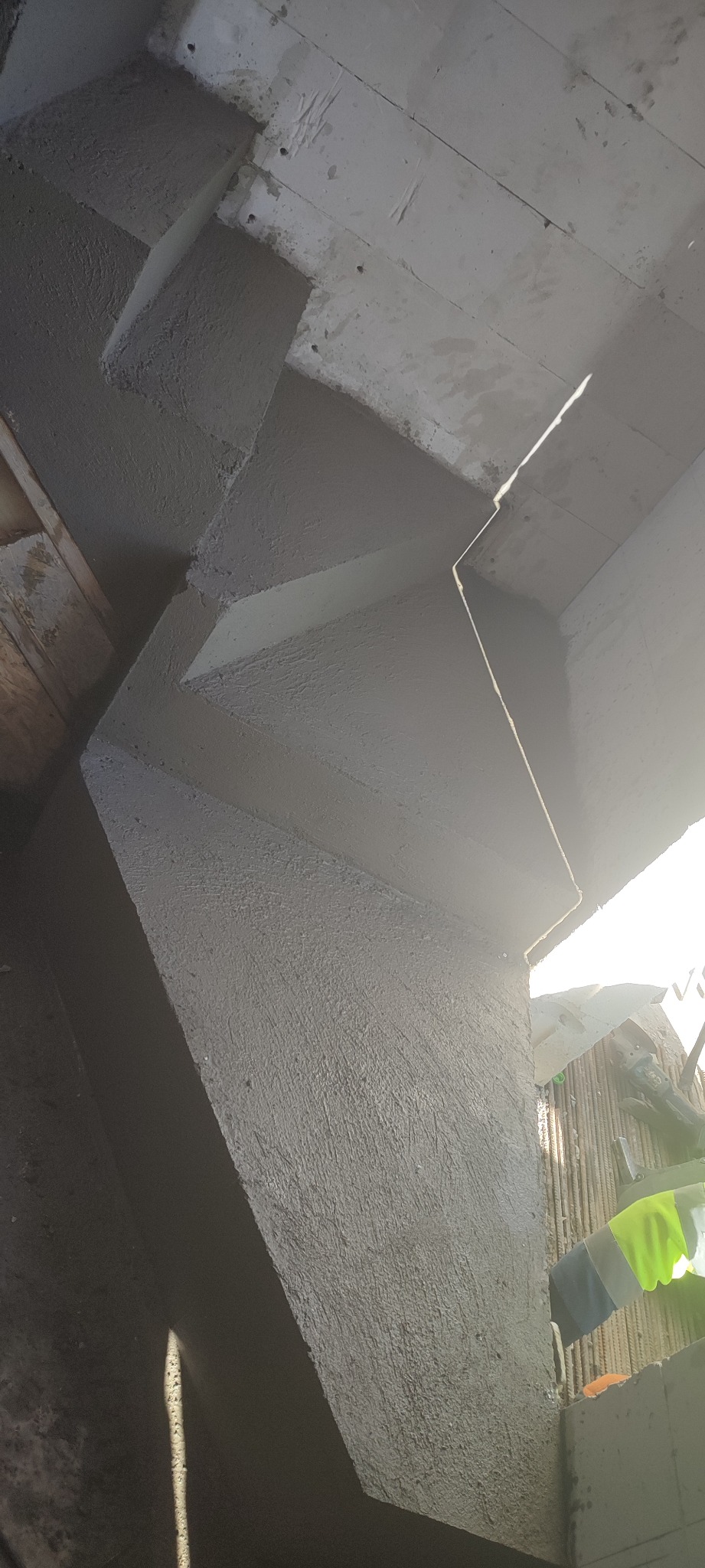 Szare, surowe ściany żelbetowe w trakcie budowy, widoczne zbrojenia, oświetlone mocnym, bocznym światłem słonecznym, tworzące geometryczne cienie na powierzchni betonu.