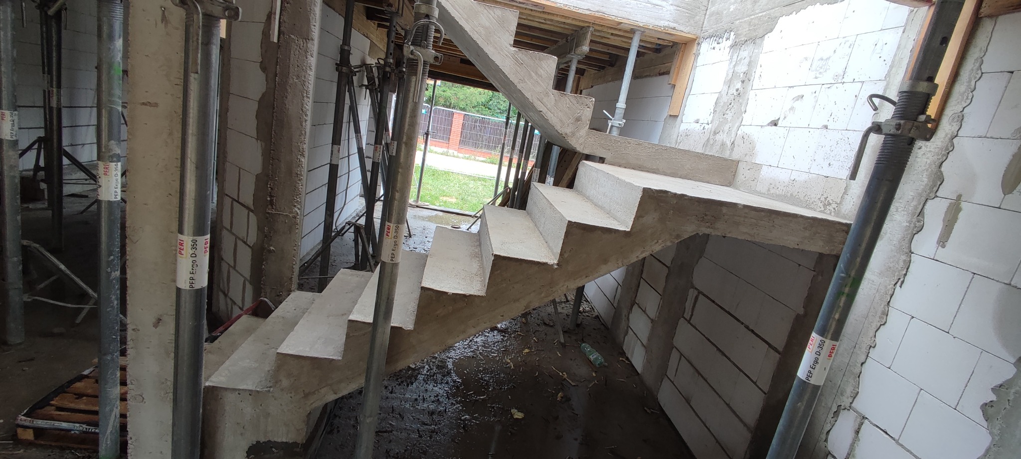 Surowe, żelbetowe schody wewnętrzne w trakcie budowy, wsparte na metalowych podporach PERI Ergo D-350, widoczne białe bloczki ścian i tymczasowe drewniane szalunki.