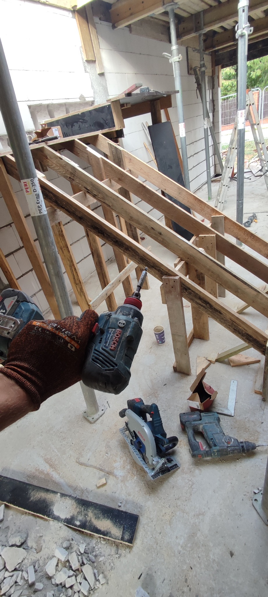 Wnętrze w trakcie budowy z widoczną konstrukcją drewnianą dachu, wspartą na stalowych podporach. W pierwszym planie ręka w roboczej rękawicy trzyma wkrętarkę Bosch, obok leżą: piła tarczowa...