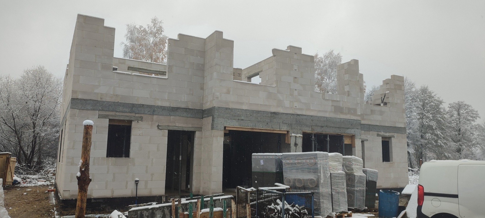 Surowy stan domu z betonu komórkowego w zimowej scenerii; widoczne przygotowane materiały budowlane i częściowo ośnieżone drzewa w tle.