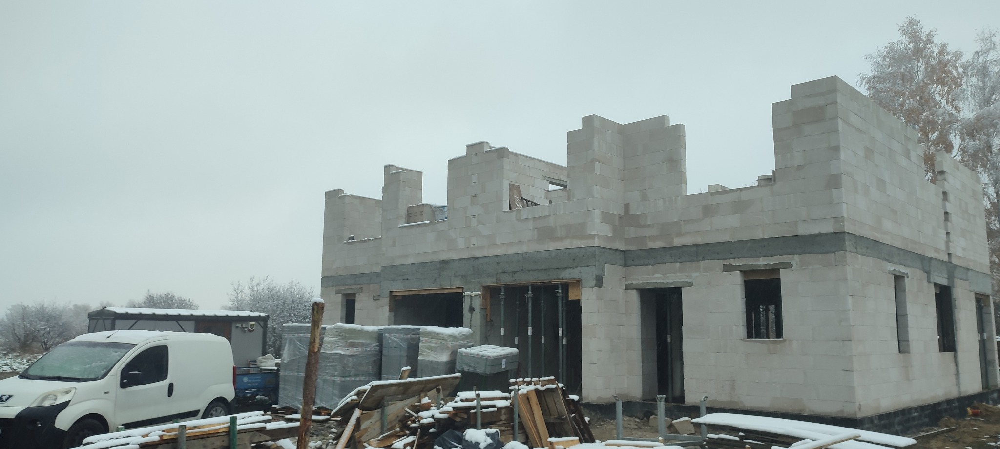 Surowy stan domu w trakcie budowy z białych bloczków, widoczne otwory okienne i drzwiowe, na pierwszym planie palety z materiałami budowlanymi pod śniegiem, w tle biały samochód dostawczy...