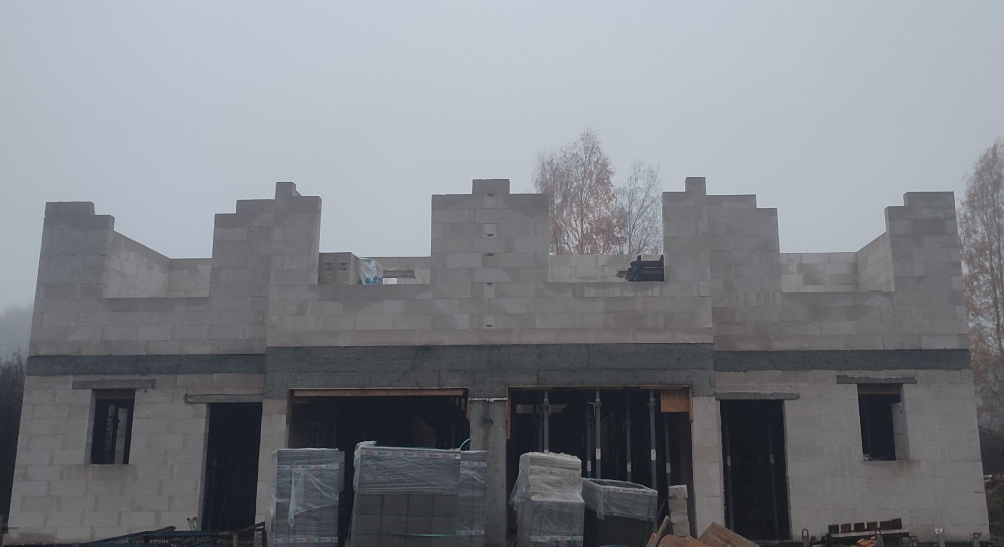 Surowy stan domu z betonu komórkowego w trakcie budowy, widoczne palety z materiałem budowlanym i zarys okien w szary, mglisty dzień.