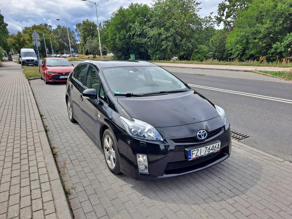 Czarny samochód osobowy Toyota Prius zaparkowany na chodniku przy ulicy, widoczny numer rejestracyjny FZI 7646A, w tle inne pojazdy i zieleń miejska.