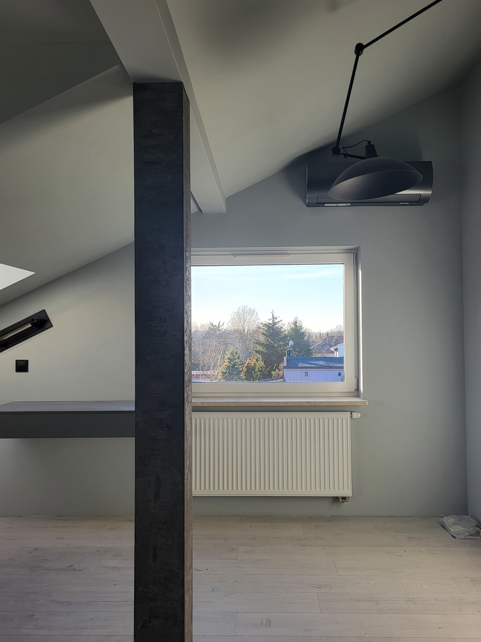 Pokój na poddaszu po remoncie, ściany pomalowane na szaro, widoczne okno z widokiem na drzewa, grzejnik, lampa sufitowa na wysięgniku, kolumna dekoracyjna z efektem betonu i jasna podłoga z paneli.
